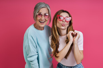 Playful mother and adult daughter wearing funky eyeglasses while standing together against pink background