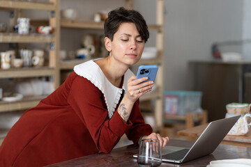 Young woman ceramic artisan using social media to communicate with customers while standing at desk with laptop in pottery studio or workshop. Craft business owner with mobile phone in ceramics shop