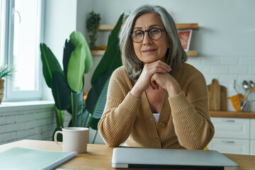 Confident senior woman looking at camera while sitting at the kitchen island at home