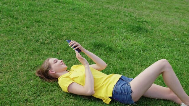 Girl in shorts lies on grass, smiles, takes selfie on smartphone. Young woman posing for cellphone pictures on lawn in summer. Female student shooting herself looking to mobile phone camera on meadow