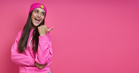 Beautiful woman in funky hat pointing away while standing against pink background