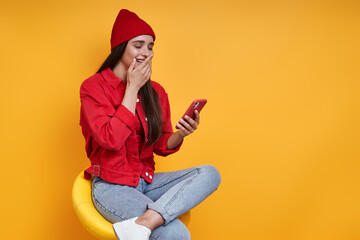 Surprised young woman using smart phone while sitting on the chair against yellow background