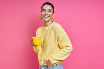 Beautiful young woman in yellow sweater holding coffee cup and smiling against colored background