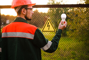 an electrician in a work uniform and a protective helmet holds a light bulb to connect lighting