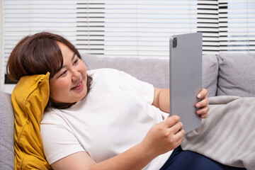 Young chubby Asian women watching online on demand movie or playing online game on digital tablet while lie down on sofa couch. With excited and happy facial expression.