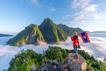 Asian girl holding Laos flag on top of Viewpoint of Nong Khiaw - a secret village in Laos. Stunning scenery of limestone cliff valley covered with fog. - 520369431