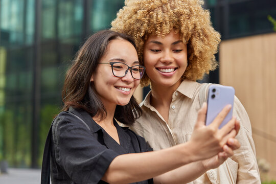 Pretty cheerful multiracial female friends take selfie together while strolling outdoors look gladfully at front camera of cellphone enjoy free time dressed in casual shirts. Friendship concept
