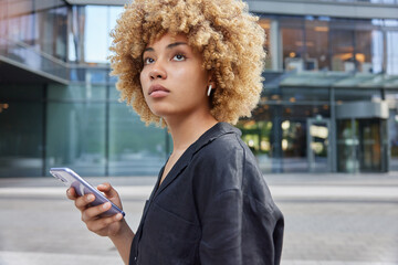 Young curly haired woman holds smartphone for blogging enjoys leisure time dressed in casual black shirt strolls against urban background focused into distance. Cellular technology lifestyle concept