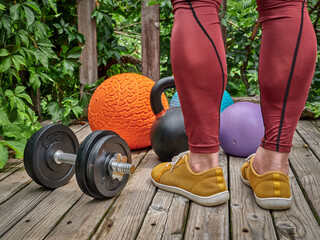 male in compression pants is exercising with heavy slam balls, dumbbell and kettlebell on a backyard deck, functional fitness and backyard gym concept