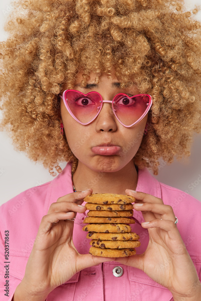 Wall mural Vertical shot of unhappy young woman with curly bushy hair holds delicious cookies feels temptation to eat sweet food wears sunglasses and stylish jacket poses indoors. Tasty homemade snack.