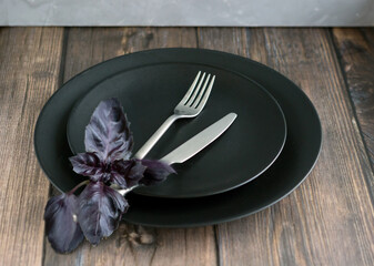 Table setting with black plate, silver cutlery with basil leaves on the wooden table.