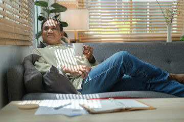 Happy senior man relaxing on couch and drinking coffee, looking out the window. Retirement lifestyle.