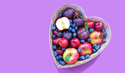 Red apples, blueberry and plums in straw basket made in the shape of heart on light shade of lavender background. Creative copy space