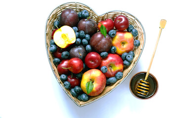 Fruit and honey for the Jewish New Year. Red apples and plums in straw basket made in the shape of heart on white background.