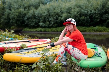 The woman sits in anticipation, sad. Beach on the small river. Girl surfer in red t-shirt sitting...