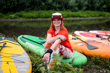 Happy smile woman surfer in red t-shirt sitting on the sup board. Beach on the small river....