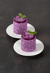 Blueberry mousse cream dessert of cylindrical shape with curly jelly on top, sprinkled with colored coconut shavings, on a plate. Dark grey background