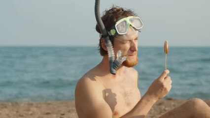 A man with a red beard on the beach, wearing scuba goggles and with a pipe, licks a multi-colored lollipop and looks at it. The sea is in the background. slow mo