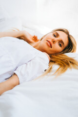 Portrait of attractive caucasian happy woman with blonde hair, lying on a bed with white blanket on sunny day.