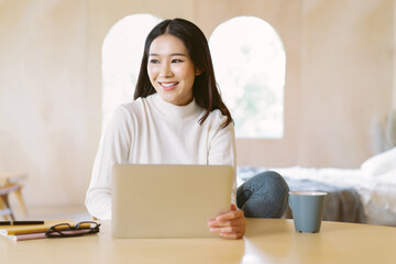 Beautiful smiling freelancer Asian woman working remotely on laptop and looking away in home winter...