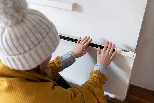 A woman in warm clothes holds her hand on the heater. Low heating temperature in the house. Energy crisis concept