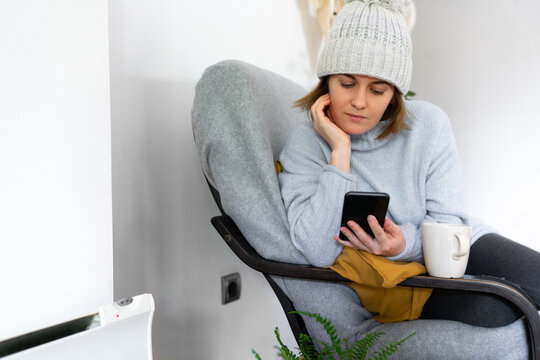 A woman in warm clothes with smartphone sits close to a heating radiator. Low temperature in the house. Energy crisis concept