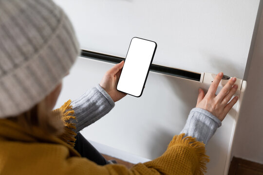 Woman's hand on heater thermostat. In the second hand is a smartphone with a white screen. Low heating temperature in the house. Energy crisis concept