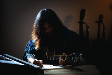 Asian musician who have long hair and wearing jean jacket playing guitar and writing a song in...
