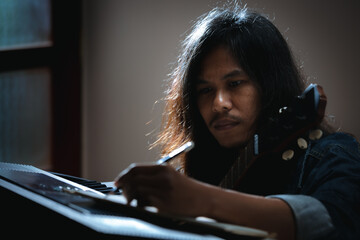 Asian musician who have long hair and wearing jean jacket playing keyboard and guitar and writing a...