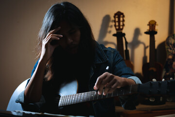 Asian musician who have long hair and wear jean jacket is playing guitar and so stressed to think about lyric and write a song in notebooks in the dark room.