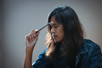 Obraz premium Asian musician who have long hair and wear jean jacket is playing guitar and so stressed to think about lyric and write a song in notebooks in the dark room.