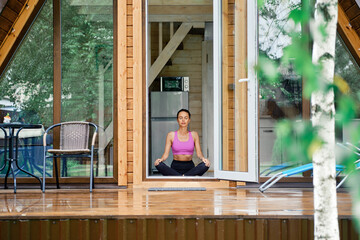 Woman sits in yoga lotus position in forest house in front of opened door