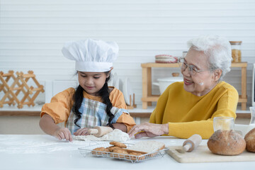 Happy family Asian elderly grandmother and little cute grandchild spend time together at kitchen, knead dough by rolling pin and bake cookies or bread, smile and have fun, flour mess up on face