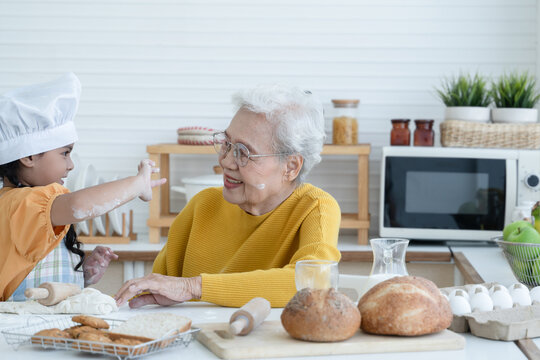 Happy family Asian grandmother and little grandchild spend time together at kitchen, knead dough and bake cookies or bread, niece have fun to tease and put flour on her grandma face at home