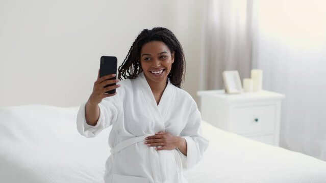 Young happy african american woman woman video chatting with friends via smartphone, sitting on bed at home, free space