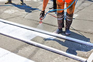 A road worker paints a white road marking of a pedestrian crossing using a wooden frame template...