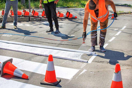 A team of road workers paints a white road marking of a pedestrian crossing using a wooden frame template and an airbrush.