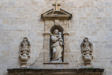 Detail of the facade of the ancient dominican convent with stone statue of the Virgin Mary holding baby Jesus in niche surmounted by cross in the historic center of Montpellier, France 