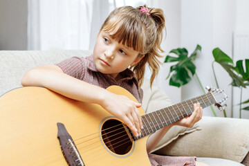 A cute little girl is sitting on the couch at home and learning to play the acoustic guitar. Hobbies for children. Learning to play a musical instrument