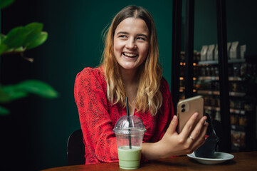 Young woman drinking green drink ice matcha latte in cafe and using smartphone.