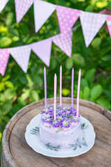 Very beautiful bento cake with purple, veri peri, matthiola flowers with green leaves, candles in the cake on the background of pink flags. Birthday. Vertical photo.