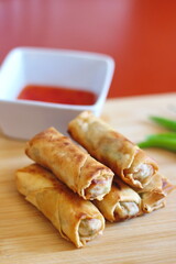 Fried chinese spring rolls placed on a kitchen chopping board and served with chilli sauce and fresh chili.