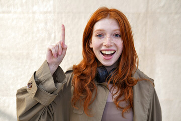 Happy teen redhead fashion girl standing on urban wall background looking at camera pointing finger up. Excited teenage girl with red hair having great new idea concept, outdoors.