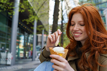 Smiling cool teenage hipster girl eating ice cream on big city urban street. Pretty teen generation z girl with red hair enjoying delicious tasty icecream spending time in park outdoors. - Powered by Adobe