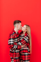 Happy Saint Valentine's Day. Romantic young couple in the same pyjamas is kissing on red background. Man and woman in love are enjoying spending time together while celebrating, copy space for text.