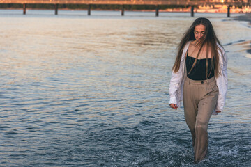 A woman walks along the sea barefoot at sunset, copy space.