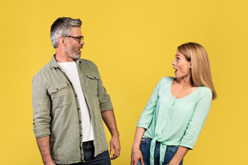 Shocked middle aged woman and her husband looking at each other with open mouths, feeling surprised on yellow background