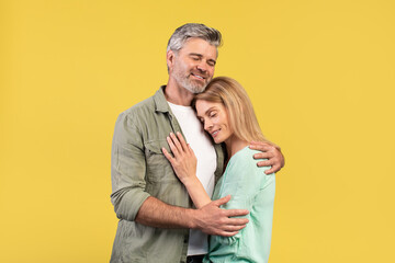 Portrait of loving middle aged spouses hugging with closed eyes while standing isolated over yellow...