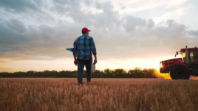 Agriculture. Working farmer walks with tablet across field against background of tractor. A worker on tractor cultivates and fertilizes field. Farmer controls operation of tractor in the wheat field.