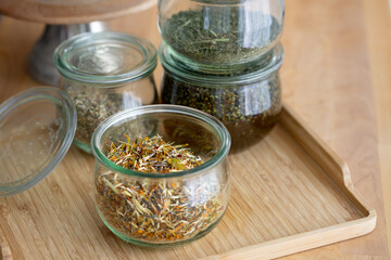Glass jars with dried flowers and herbs stand on wooden table with light reflections on the jars, homemade aroma oil ingredients